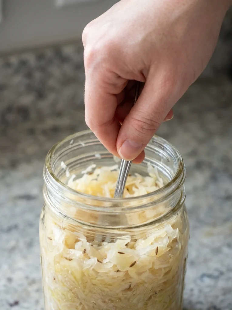 A hand using a fork to mix sauerkraut in a glass jar.